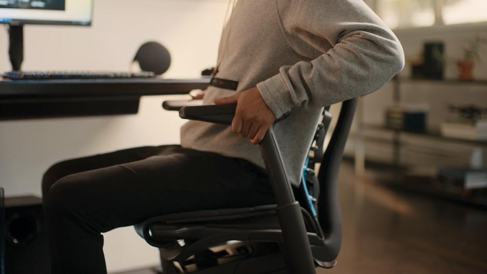 En video af en person, der sidder i en sort og blå Embody-gamingstol fra Herman Miller og rækker ud efter stolen for at justere armene.
