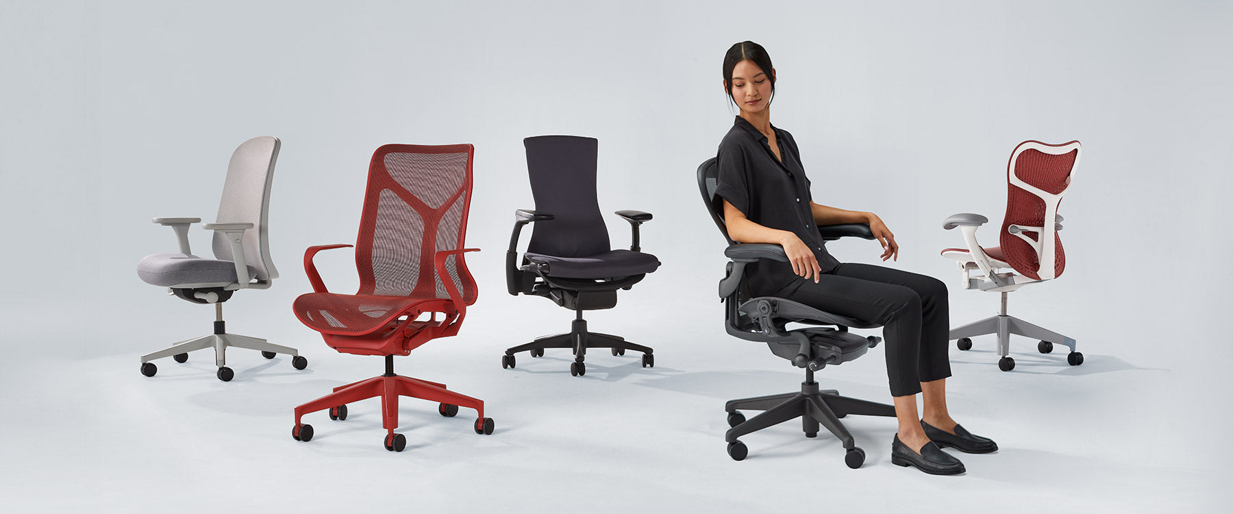 Foto af Herman Miller-stole på række, herunder Verus mineralhvid og grå, Cosm med mellemhøj ryg i canyonrød, Embody sort, Aeron grafit med dame siddende og Mirra 2 i cordovanrød