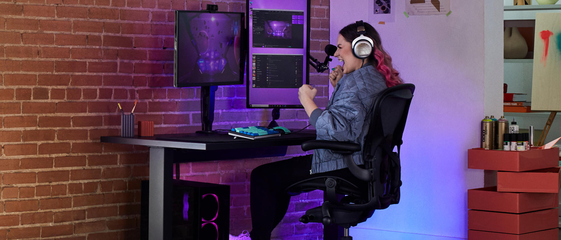 Electra smiler med knyttede næver, mens hun gamer siddende på Aeron gaming onyx black med Nevi gaming-skrivebord i sort og Ollin-skærmarm