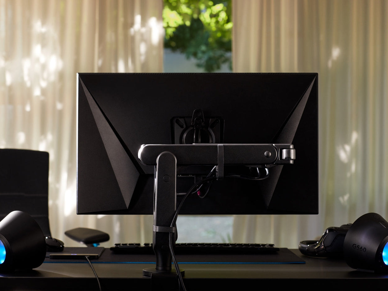 Bagfra ses en sort Ollin-skærmarm, der holder en computerskærm og er fastgjort til et sort Nevi-gamingskrivebord fra Herman Miller. Set i et spillerum eller hjemmekontor, med lys, tastatur og mus.