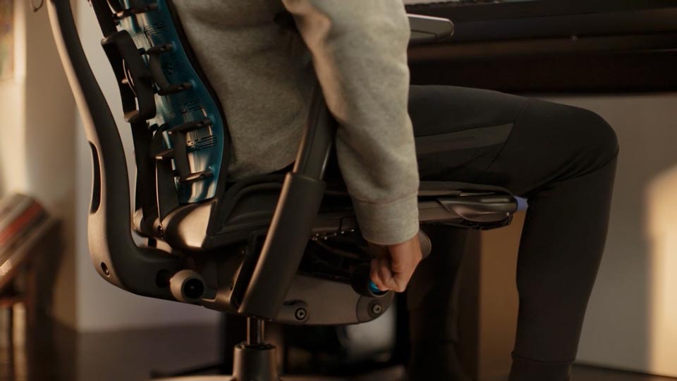 En video af en person, der sidder i en sort og blå Embody-gamingstol fra Herman Miller og rækker ud ved siden af stolen for at justere sædehøjden.