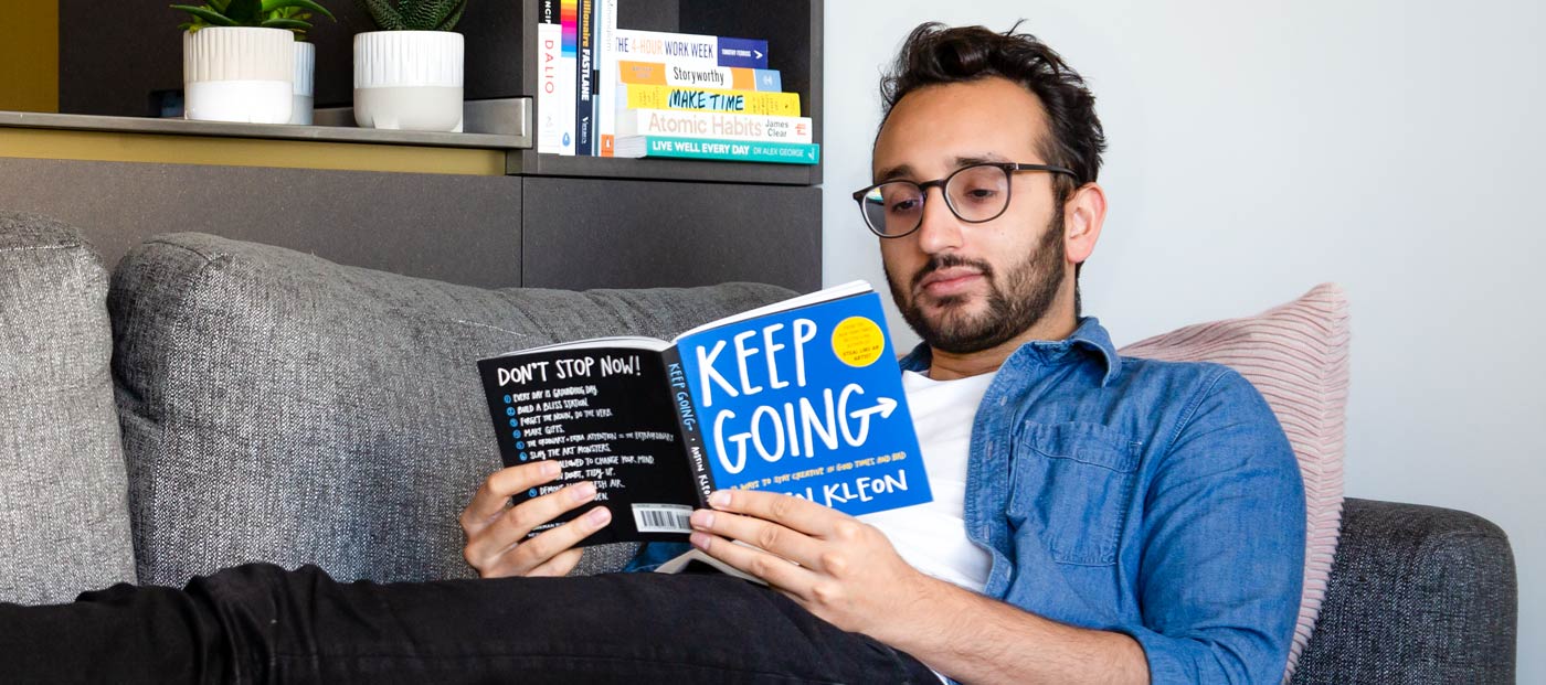 Foto af Ali Abdaal liggende på sofaen og læsende en bog ved navn Keep Going