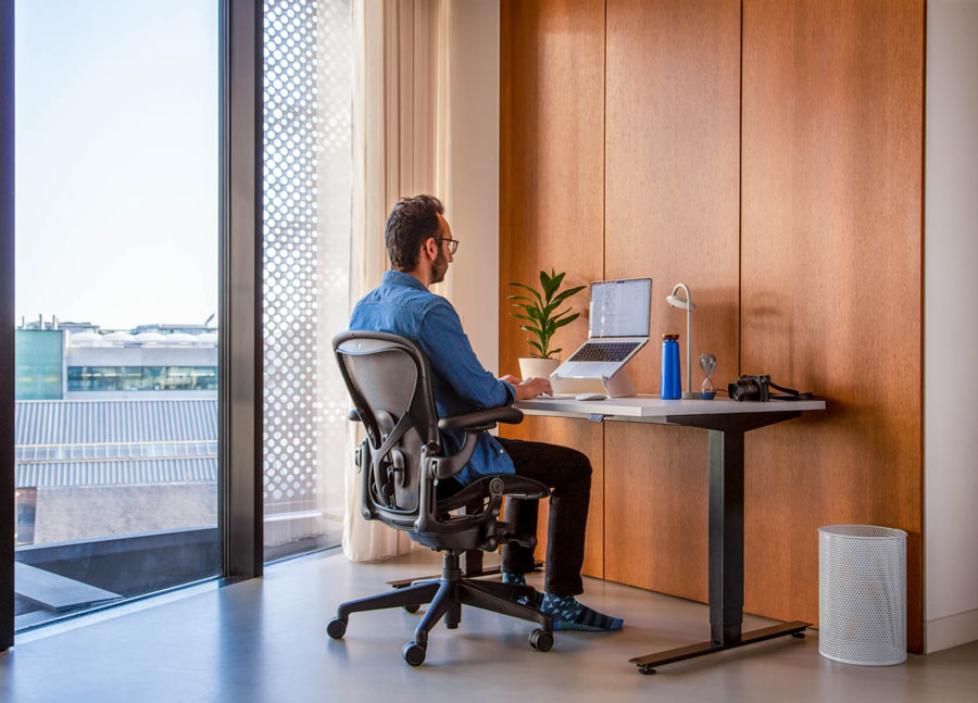 Foto af Ali Abdaal, podcaster, siddende i en Herman Miller Aeron-kontorstol i grafit foran et Nevi-siddestativ med sorte ben og hvid bordplade med en bærbar computer i en Oripura-laptopholder