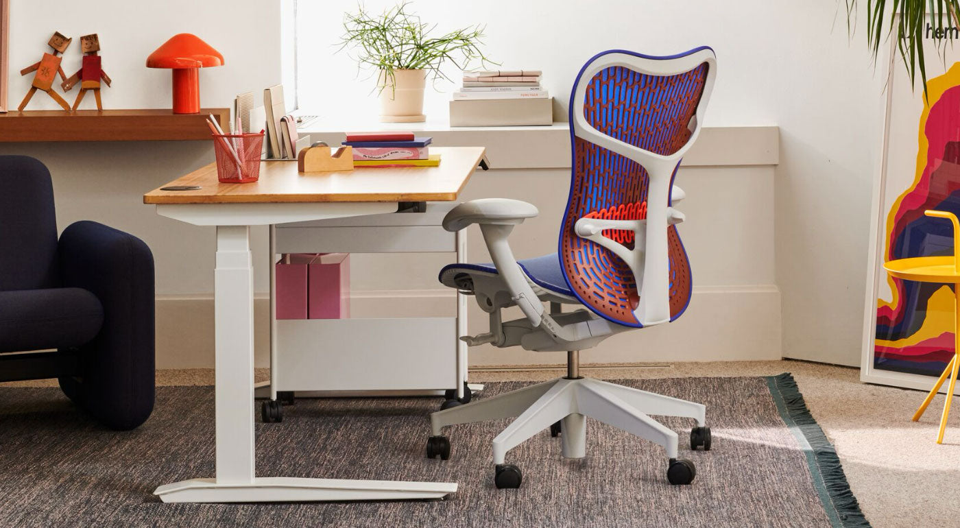 En blå, orange og hvid Mirra 2-kontorstol fra Herman Miller, placeret ved siden af et Jarvis-skrivebord. Opstillet i et hjemmekontormiljø med en blå Wilkes-sofa, et blåt Maharam-tæppe, et gult DLM-sidebord og en rød HAY Pao-bordlampe.