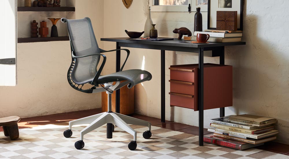 Et sort Mode-skrivebord fra Herman Miller med opbevaring i terracotta og håndtag i valnød er placeret foran en grå Setu-kontorstol fra Herman Miller. Set i et velindrettet hjemmekontor med hylder, bøger og dekorativt tilbehør til hjemmet.