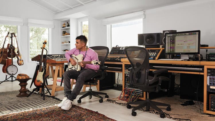 Foto af Rostam, musiker, producer og komponist, i sit musikstudie, hvor han spiller guitar siddende i en Aeron-kontorstol i grafit