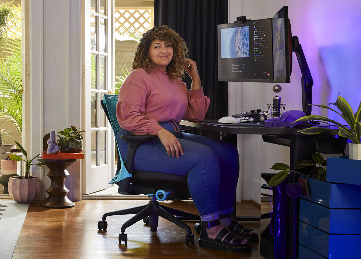 En kvinde sidder i en blå og sort Sayl-gamingstol fra Herman Miller, placeret bag et sort Nevi-skrivebord med en sort Ollin-skærmarm og -skærm. Set i et indendørs rum med trægulve, hvide vægge og indendørs planter.