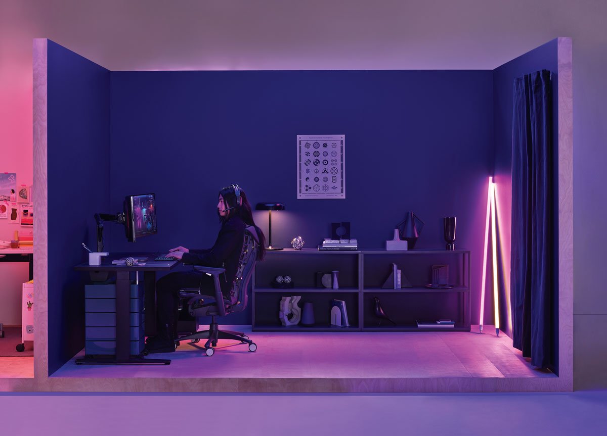 Gaming-kontor med kvinde, der spiller, siddende i en Logitech Embody-gamingstol i sort med Nevi-gamingbord i sort og Hay-neonrørbelysning i hjørnet