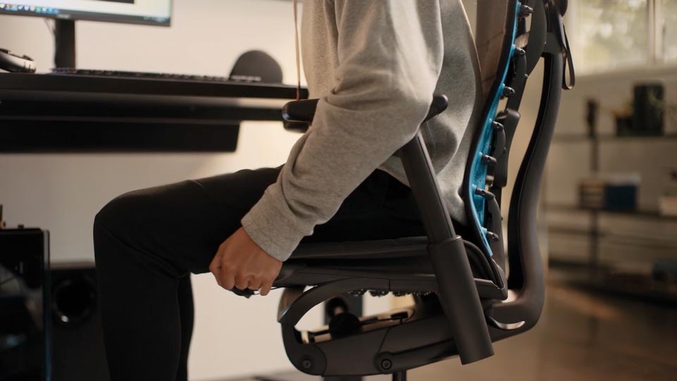 En video af en person, der sidder i en sort og blå Embody-gamingstol fra Herman Miller, og rækker ud efter stolen for at justere sædedybden.