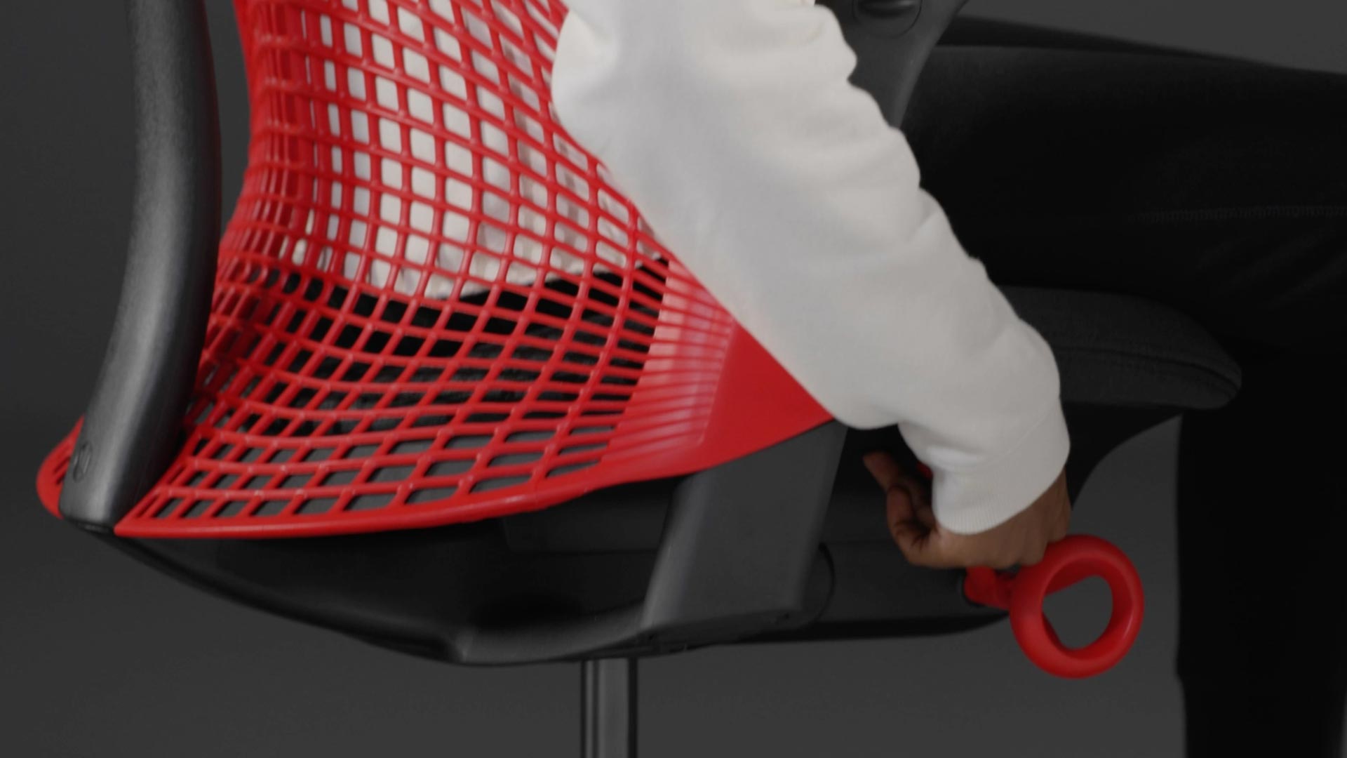 En video af en person, der sidder i en rød og sort Sayl-gamingstol fra Herman Miller og rækker ud efter stolen for at justere sædehøjden.