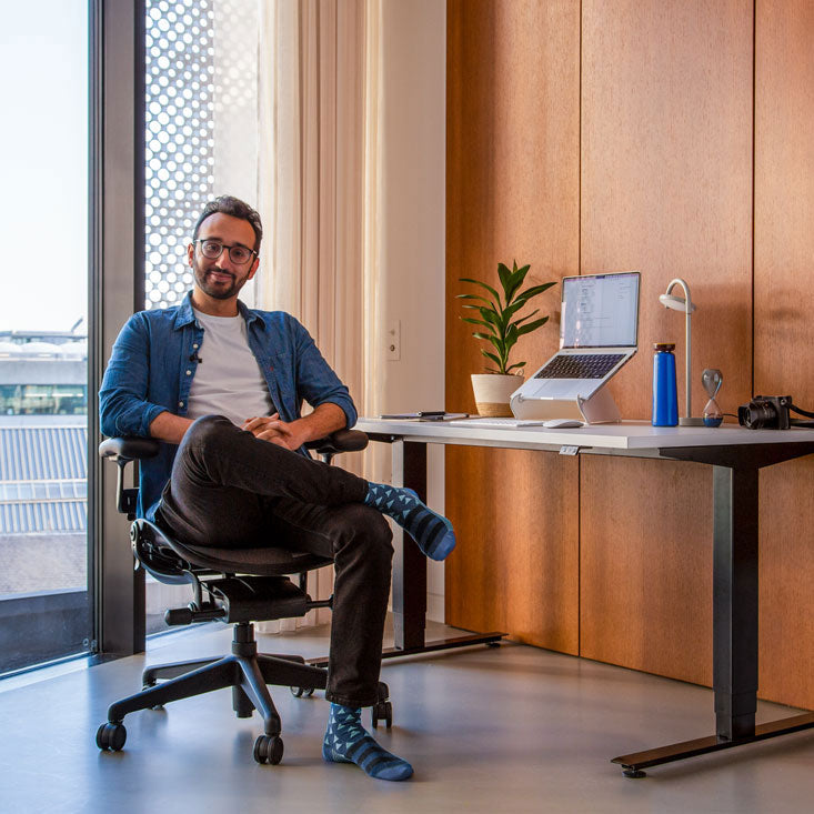 Foto af Ali Abdaal, podcaster, siddende i en Herman Miller Aeron-kontorstol i grafit foran et Nevi-siddestativ med sorte ben og hvid bordplade med en bærbar computer i en Oripura-laptopstativ