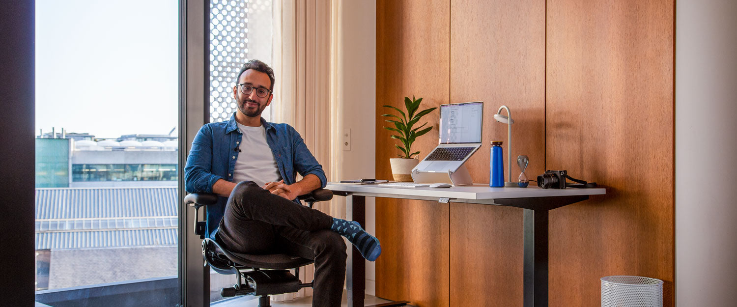 Foto af Ali Abdaal, podcaster, siddende i Herman Miller Aeron kontorstol i grafit foran Nevi sit-stand skrivebord med sorte ben og hvid bordplade med en bærbar computer i en Oripura laptop-holder