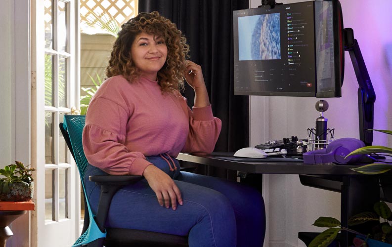 Pro streamer og stemmeskuespiller Friskk siddende på Herman Miller Sayl gaming-stol i Ocean deep blue og sort ved et sort Nevi sit-stand gaming-bord med skærm på Oliin-skærmarm.
