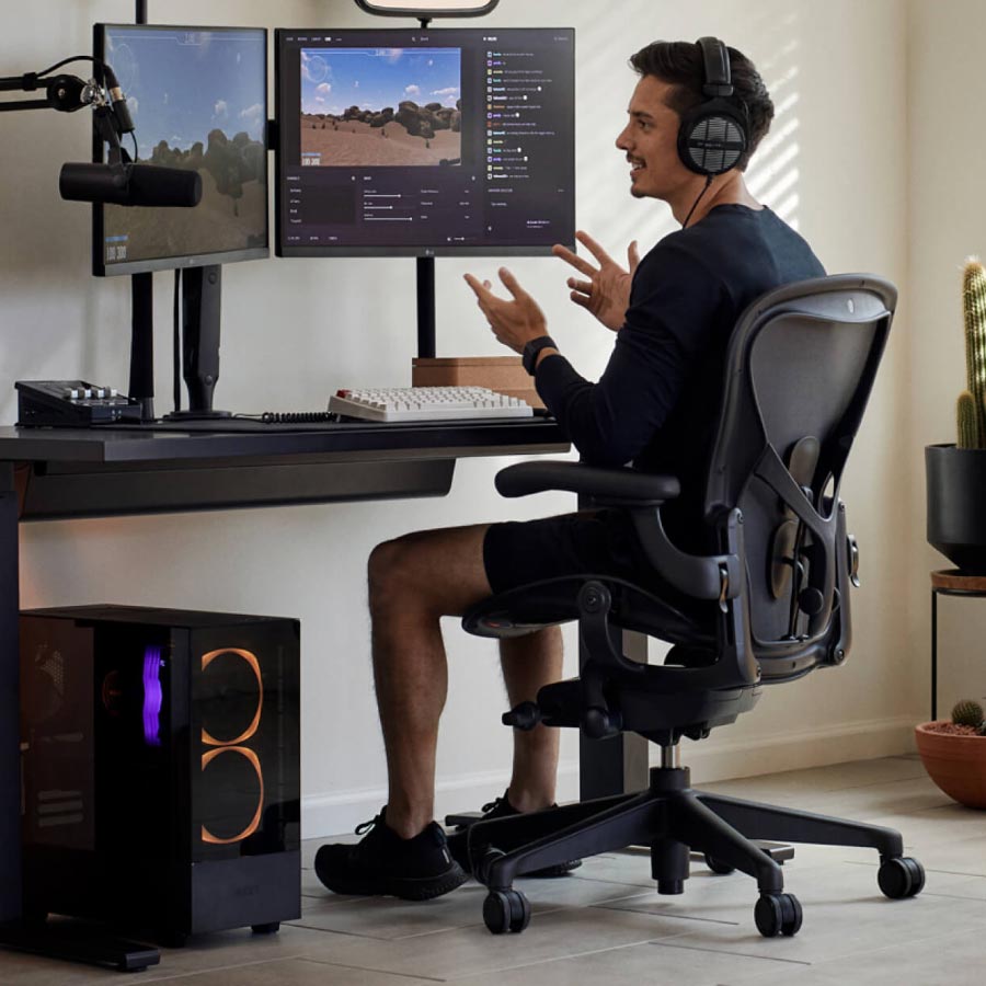 Gamer og streamer Xaryu i en gaming-opsætning siddende på Herman Miller Aeron gaming-stol med sort Nevi sit-stand gaming-bord og dobbelte skærme på Ollin-skærmarme