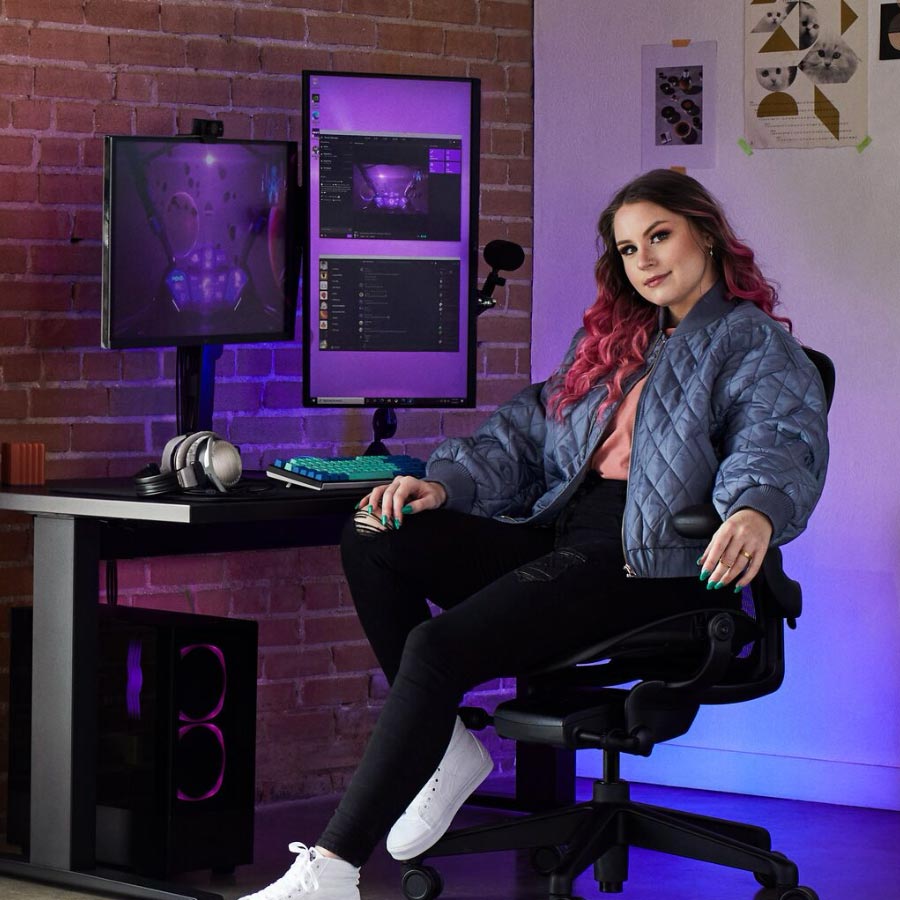 Stjernespiller og coach Electra sidder ved et gaming-set med Herman Miller Aeron gaming-stol og Nevi sit-stand-bord i sort med to skærme på Ollin-skærmarme