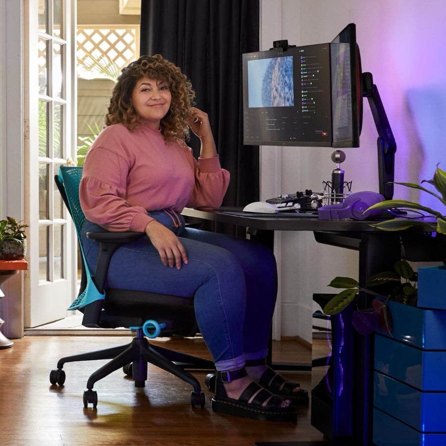 Pro streamer og stemmeskuespiller Friskk sad på Herman Miller Sayl gaming-stol i Ocean deep blue og sort ved et sort Nevi sit-stand gaming-bord med skærm på Oliin skærmarm.