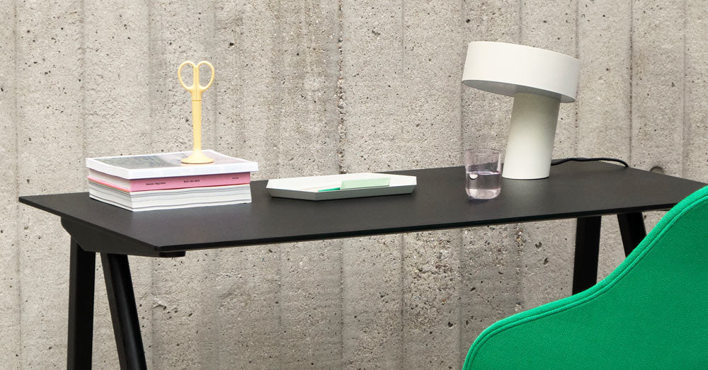 En hvid Slant-lampe fra HAY, placeret på et sort Copenhague 90-skrivebord fra HAY, sammen med en stak bøger, en gul saks og et vandglas.