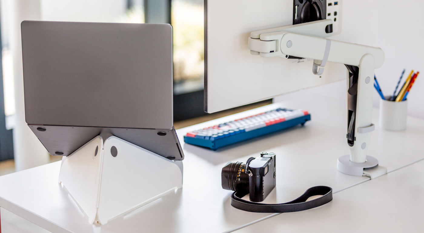 Et hvidt Oripura-stativ til bærbare computere fra Herman Miller holder en bærbar computer sammen med en hvid monitorarm.