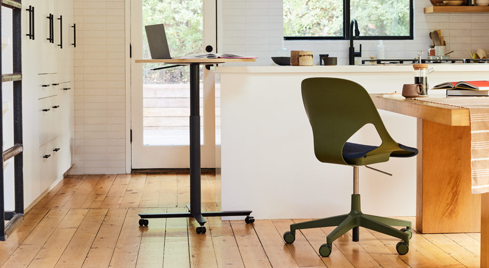 Et sort og egetræsbaseret Passport-bord fra Herman Miller placeret i nærheden af en hvid køkkenø med en bærbar computer og en bog. På billedet ses også en grøn og sort Zeph-stol fra Herman Miller placeret ved et spisebord af træ.