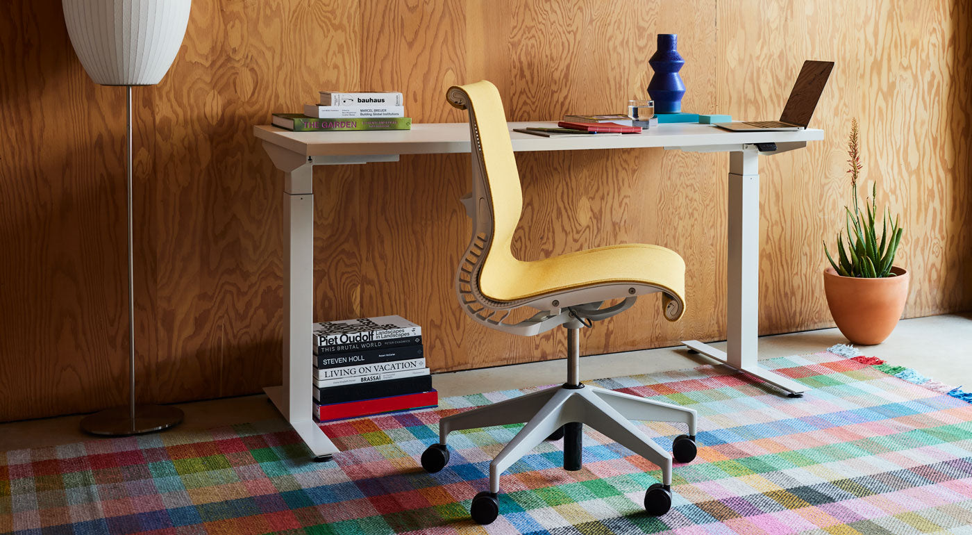 En gul Setu-kontorstol fra Herman Miller, betrukket med Mimosa Meld-stof, placeret foran et hvidt Nevi-skrivebord på et farverigt, ternet tæppe.
