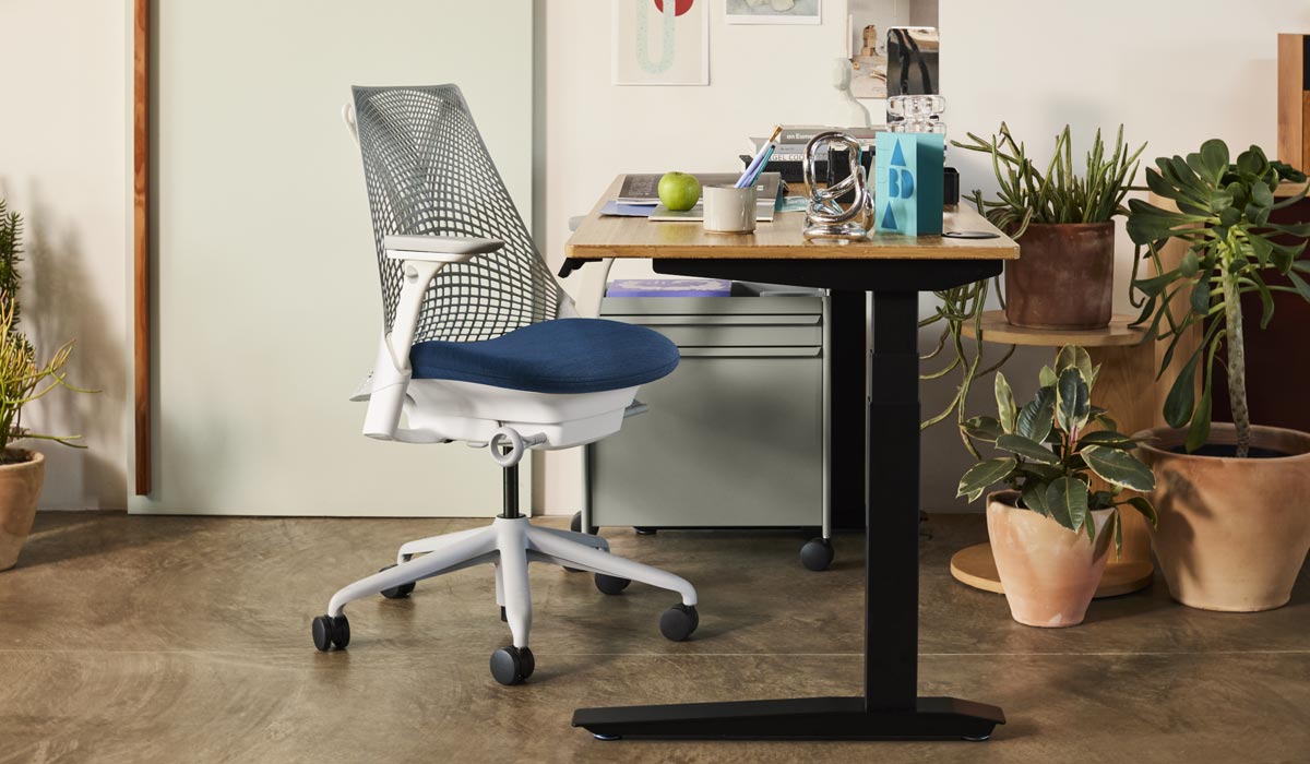 En hvid og blå Sayl-kontorstol fra Herman Miller, placeret ved siden af et Jarvis-skrivebord med sorte ben og en bordplade af bambus. I et hjemmekontormiljø med en blå OE1-trolley og et udvalg af indendørs planter i terrakottakrukker.