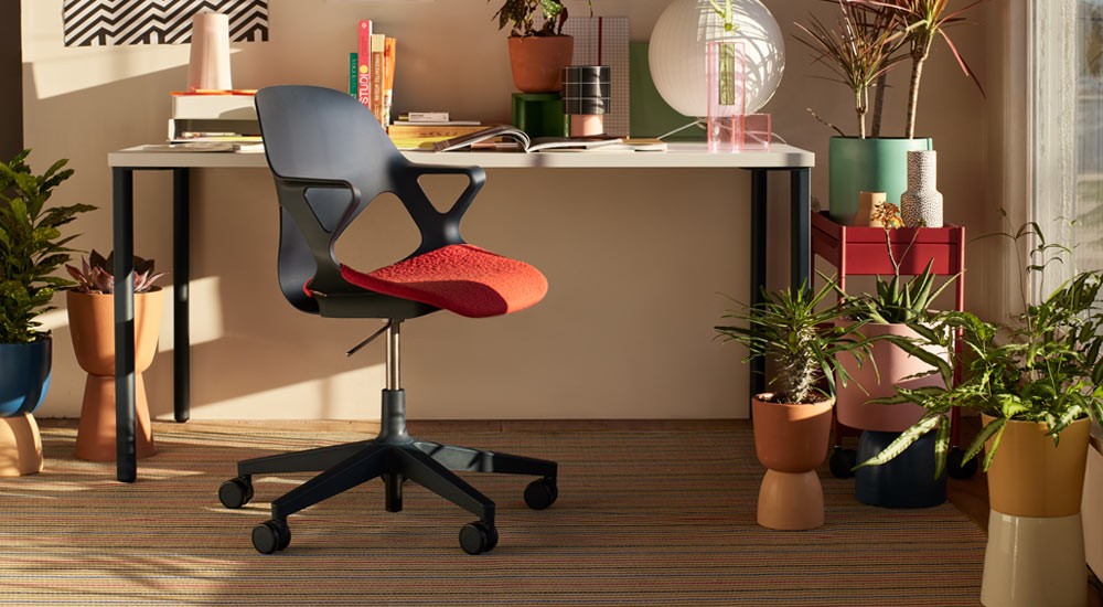 En sort Zeph-kontorstol med rød sædepude er placeret foran et Herman Miller OE1-skrivebord. Set i et hjemmekontormiljø med en rød OE1-trolley og et udvalg af indendørs planter i farverige potter.