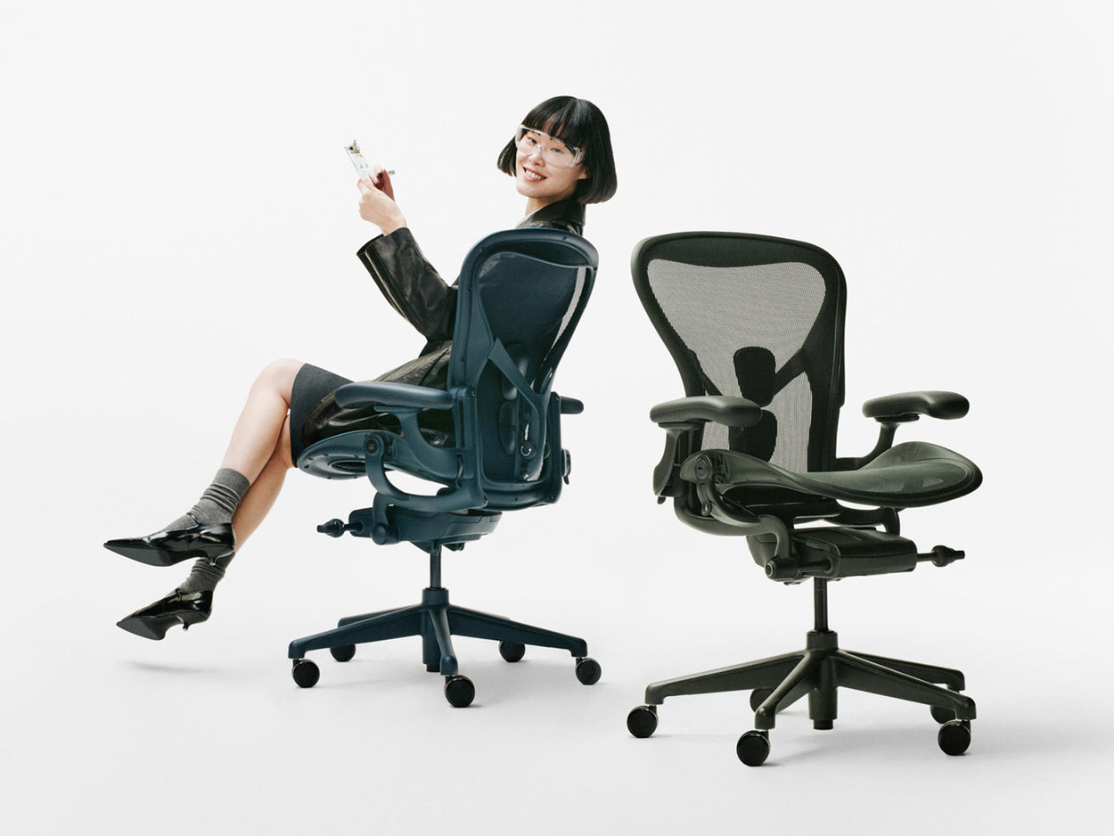 En kvinde sad på en blå Herman Miller Aeron-kontorstol i farven Nighfall ved siden af en anden Aeron-stol i farven Jasper Green i et hvidt studie.