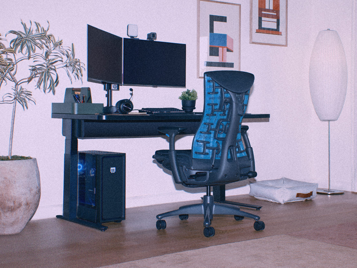 Et grynet billede af en sort og cyanblå Herman Miller Embody-gamingstol som del af et PC-gaming-setup til hjemmet, der omfatter et Nevi-gamingbord, der kan indstilles til siddende eller stående arbejde.