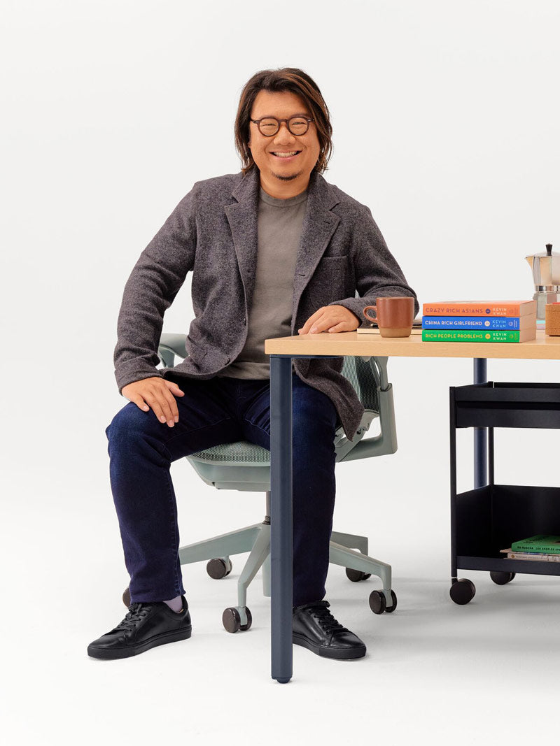 Muntert fotografi af Kevin Kwan siddende i en Glacier Grey Cosm-kontorstol, lænet mod et OE1-skrivebord med fast højde, ben i nightfall og bordplade i lys ask