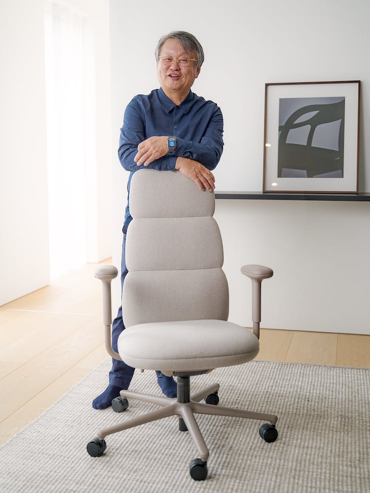 Designeren Naoto Fukasawa læner sig op ad en Herman Miller Asari-kontorstol. I baggrunden ses et rent gulv og en ren væg.