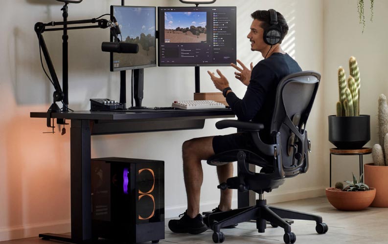 Gamer og streamer Xaryu i et gaming-setup siddende på Herman Miller Aeron gaming-stol med sort Nevi sit-stand gaming-bord og to skærme på Ollin-skærmarme