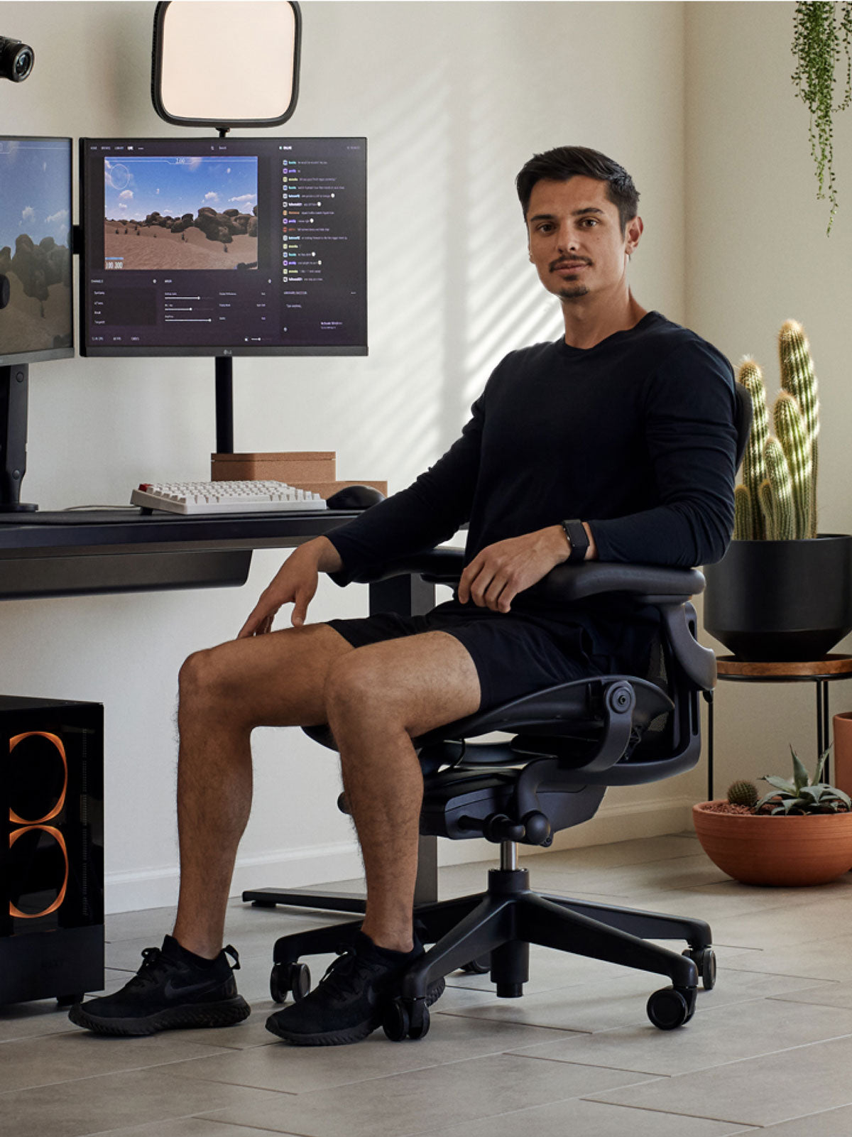 En mand sidder i en sort Aeron-gamingstol fra Herman Miller, placeret bag et sort Nevi-skrivebord med en sort Ollin-monitorarm og -skærm. I et indendørs rum med klinkegulv, hvide vægge og indendørs planter i terrakottakrukker.
