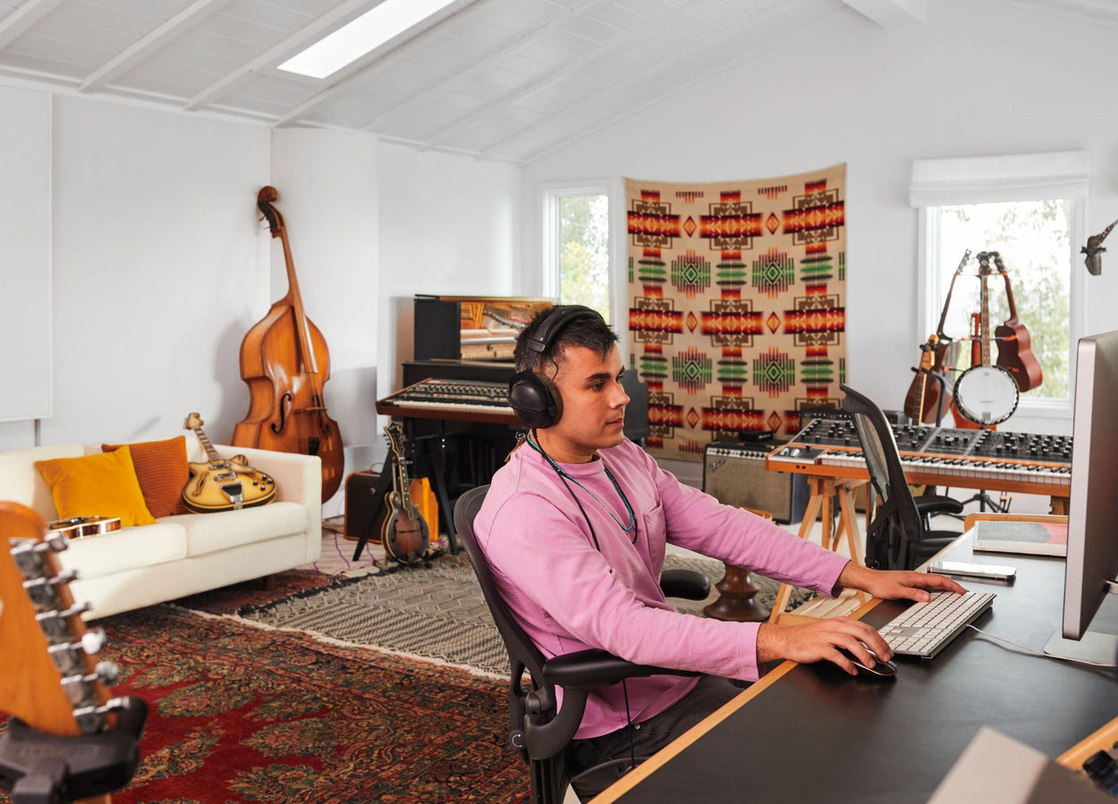 Rostam, musiker, producer og komponist, sad i en Aeron-kontorstol i grafit ved computeren med hovedtelefoner på og musikinstrumenter i baggrunden.